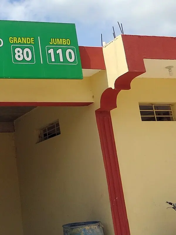 Fachada de un colmado dominicano pintada de amarillo y rojo, con su letrero verde de precios.
